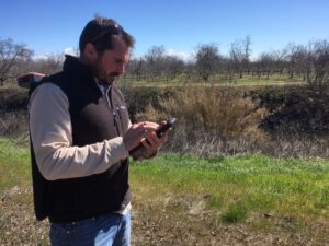 A man in a khaki shirt and dark grey vest peers down at his phone beside a river bank crowded with vegetation