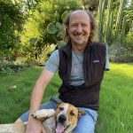 A bearded, balding man with one arm smiles. He wears a black vest, sits in a green grassy area and pets a small brown and white dog. 