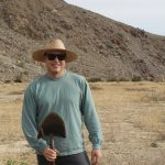 A man in sunglasses, light green long sleeve shirt and broad brimmed sunhat smiles while holding a shovel in a desert landscape by a small hill