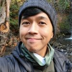 A man in a grey knit hat, neckscarf, and warm coat smiles in front of a creek and wooded area