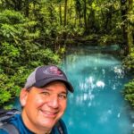 A smiling man in a baseball cap with a vivid blue lagoon and green plants in the background