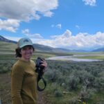 A woman in a white baseball cap and olive green long shirt holds binoculars and smiles at the camera. Behind her, low bushes and small yellow flowers dot the ground. In the distance, a river and low hills.