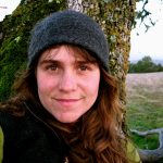 A woman with long brown hair wearing a grey knit cap smiles in front of a tree trunk